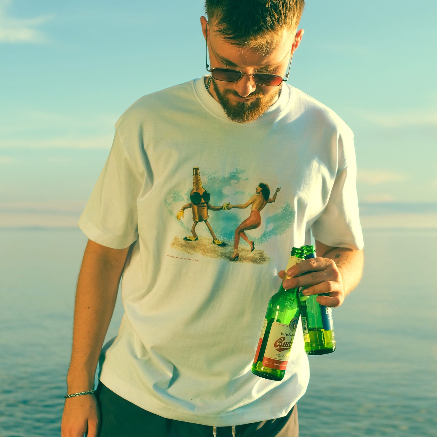 Omnia Bona ‘Dancing Beer Bottle’ Retro Graphic T-Shirt worn by a male model holding a bottle at the beach