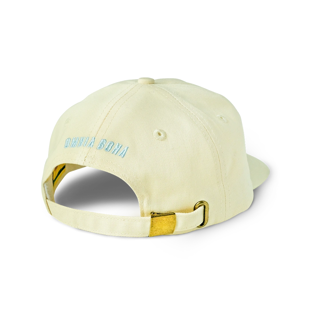 Rear view of cream-colored Omnia Bona baseball cap featuring light blue logo embroidery and gold-tone adjustable strap